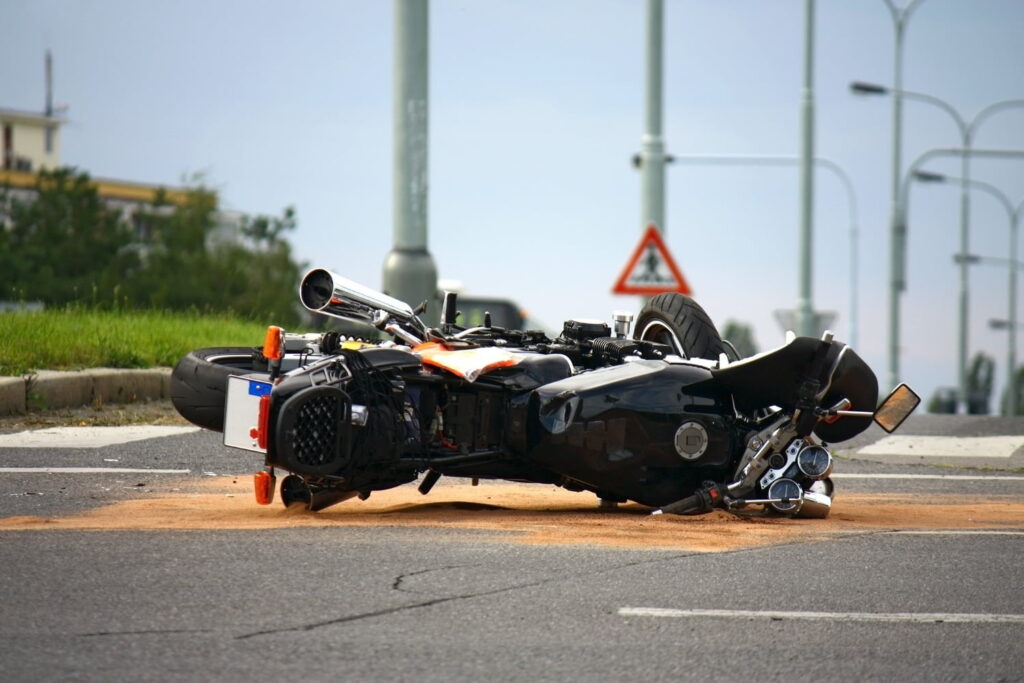 A motorcycle lies on its side on a city street, with sand underneath it, suggesting a recent accident. Traffic signs and streetlights are visible in the background.