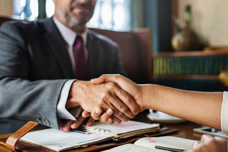 Two people are shaking hands across a desk with notebooks and papers, suggesting a business meeting or agreement.