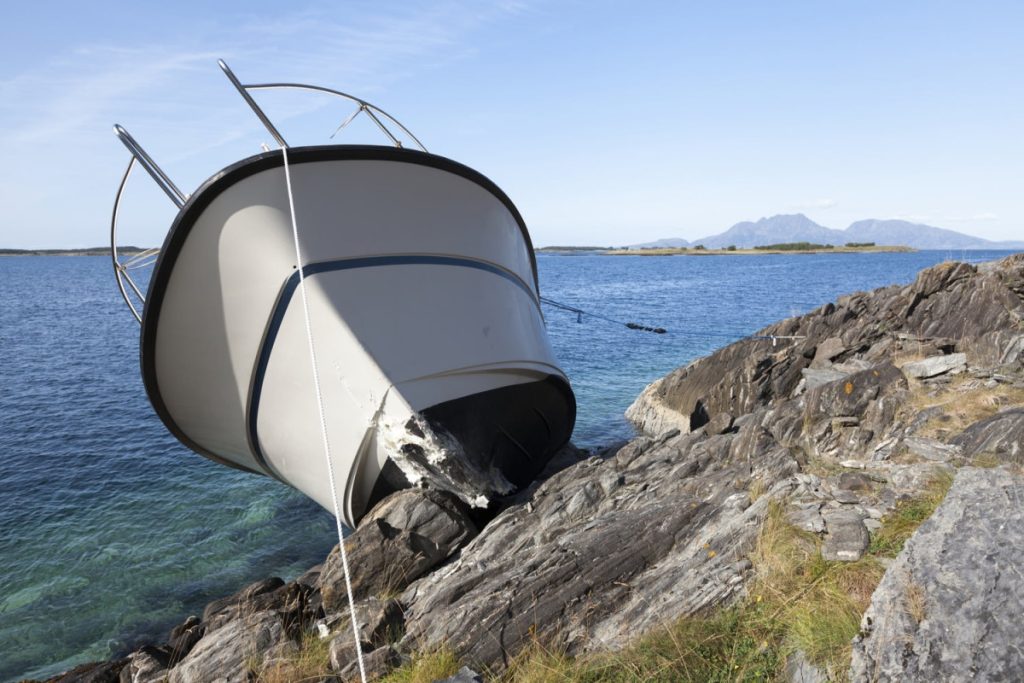A damaged boat is stranded on a rocky shoreline, with its bow resting on the rocks and the stern still in the water.