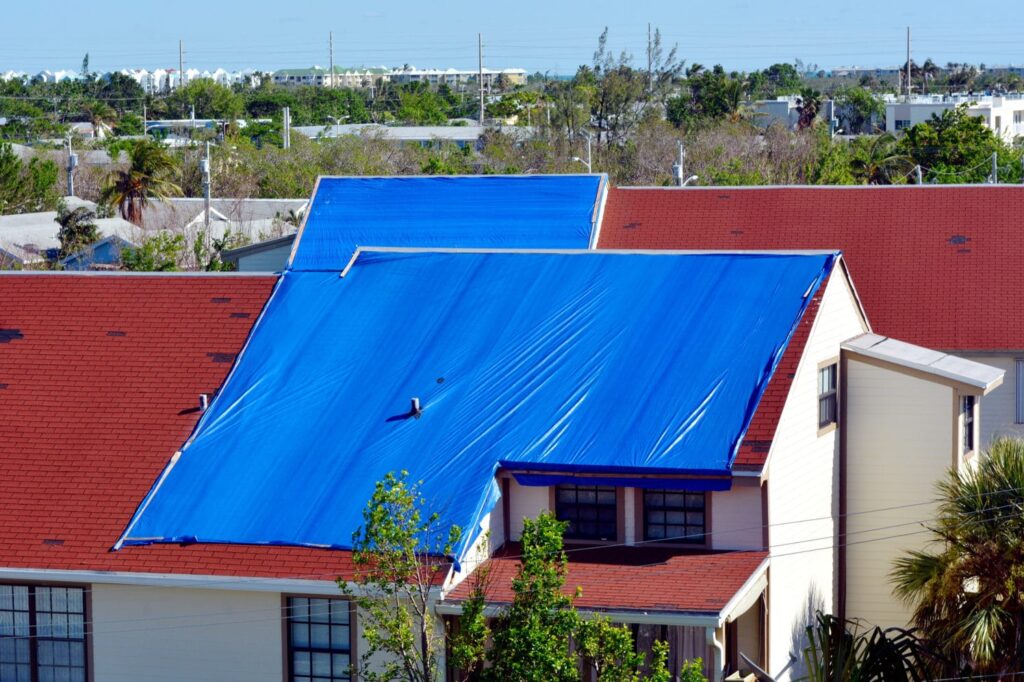 A house with a red roof has large blue tarps covering significant portions of the roof, likely for temporary weather protection or repairs.