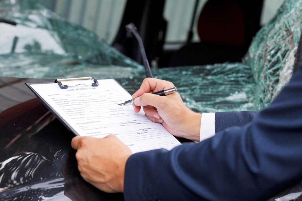 Person filling out an auto accident form on a clipboard next to a car with a shattered windshield.