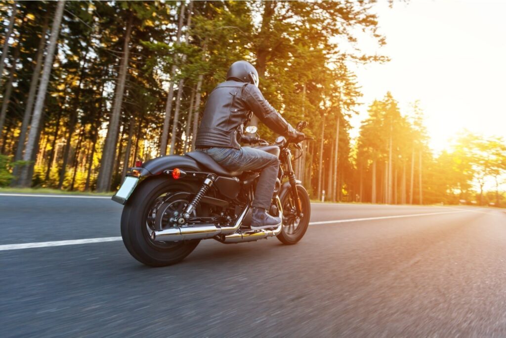 A person wearing a helmet and leather jacket rides a motorcycle on a tree-lined road at sunset.