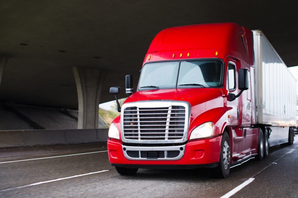 A red semi-truck with a white trailer drives on a wet highway under an overpass.