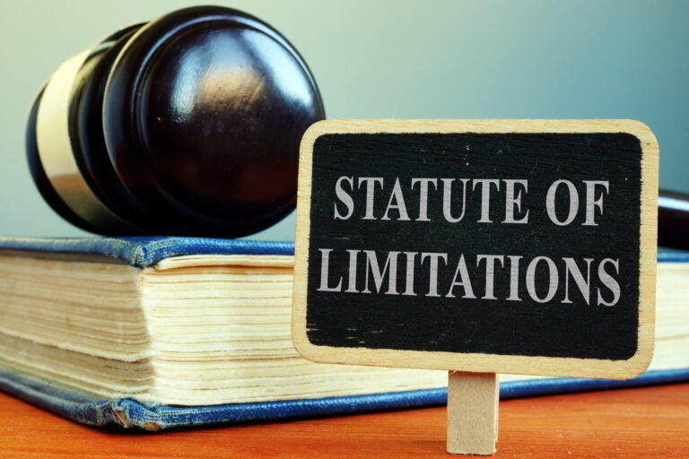 A judge’s gavel, a thick book, and a small sign reading “Statute of Limitations” are displayed on a wooden surface.