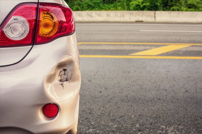 Rear bumper of a silver car with a visible dent and black scuff marks, parked on a road with yellow lines and a concrete barrier in the background.