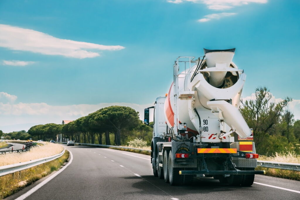 A concrete mixer truck drives on a curved, two-lane road bordered by trees and guardrails under a partly cloudy sky.