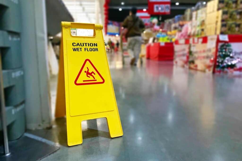 Yellow "Caution Wet Floor" sign placed on a shiny floor in a store, with blurred shoppers and shelves visible in the background.