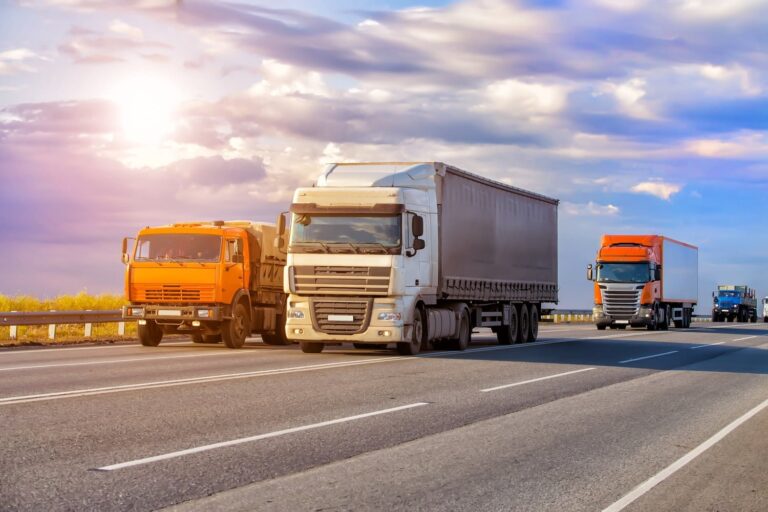 Three large trucks drive on a multi-lane highway under a partly cloudy sky, with sunlight shining in the background.