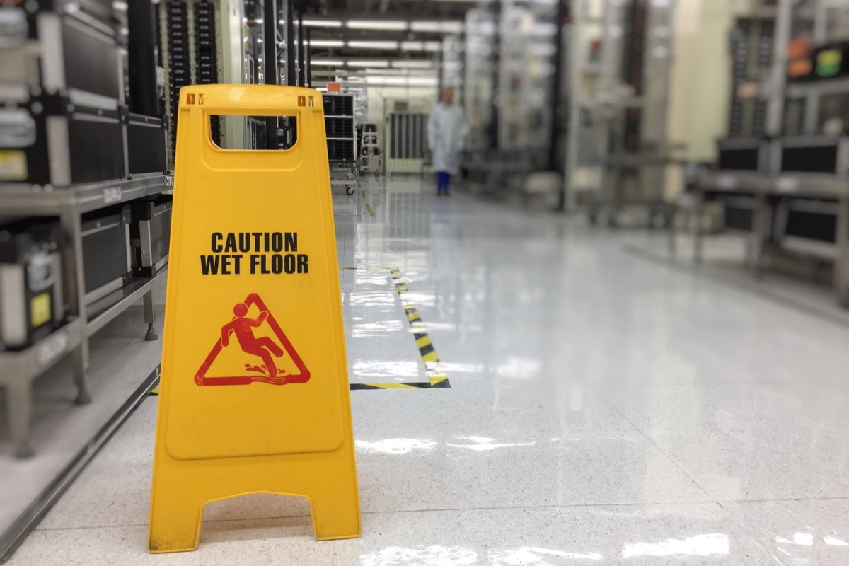 Yellow "Caution Wet Floor" sign on a shiny, reflective floor in an industrial or laboratory setting with shelves and a person in the background.