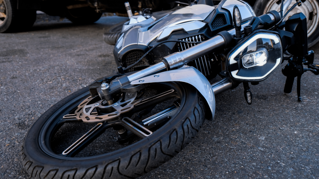 A motorcycle lies on its side on an asphalt surface, with visible front wheel, brake disc, and headlight.