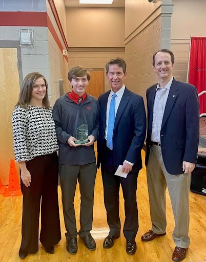 Four people stand indoors on a gym floor; one young man holds an award while the others stand beside him, all dressed in business or business-casual attire.