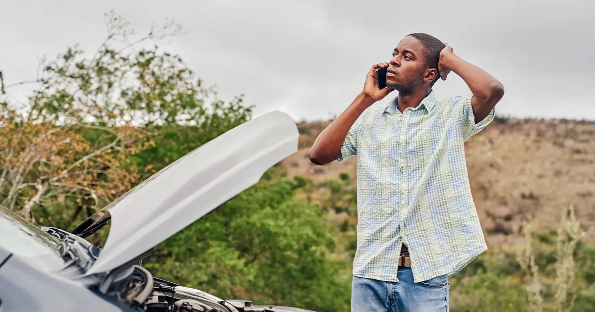 A man stands by a car with the hood open, talking on the phone and looking concerned, with trees and hills in the background.