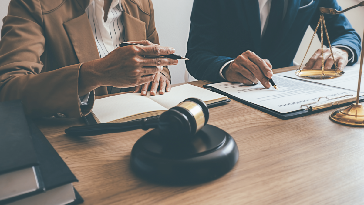 Two people in business attire discuss documents at a desk with a gavel, legal books, and scales of justice visible.