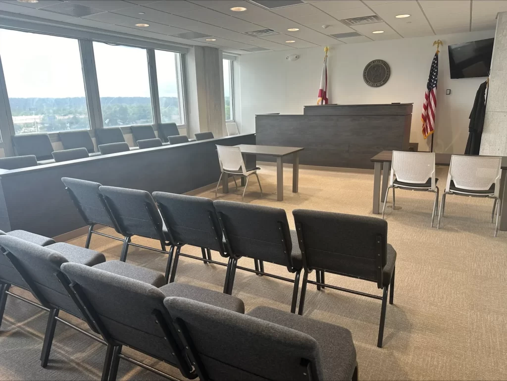 Empty courtroom with rows of gray chairs, a judge’s bench, two desks, and American and state flags in the background. Large windows let in natural light.