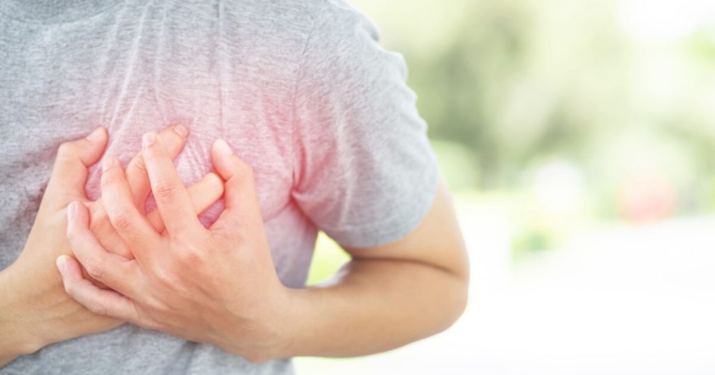A person in a gray shirt clutching their chest with both hands, suggesting chest pain or discomfort.