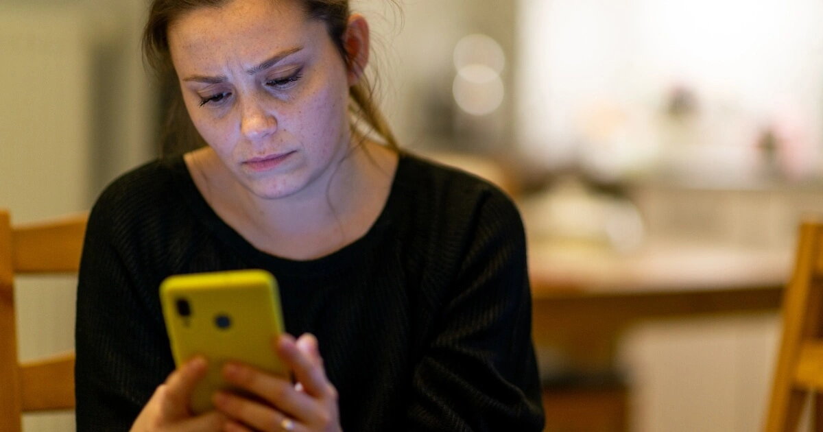 Woman sitting indoors, looking intently at a yellow smartphone with a concerned expression, in a softly lit room.