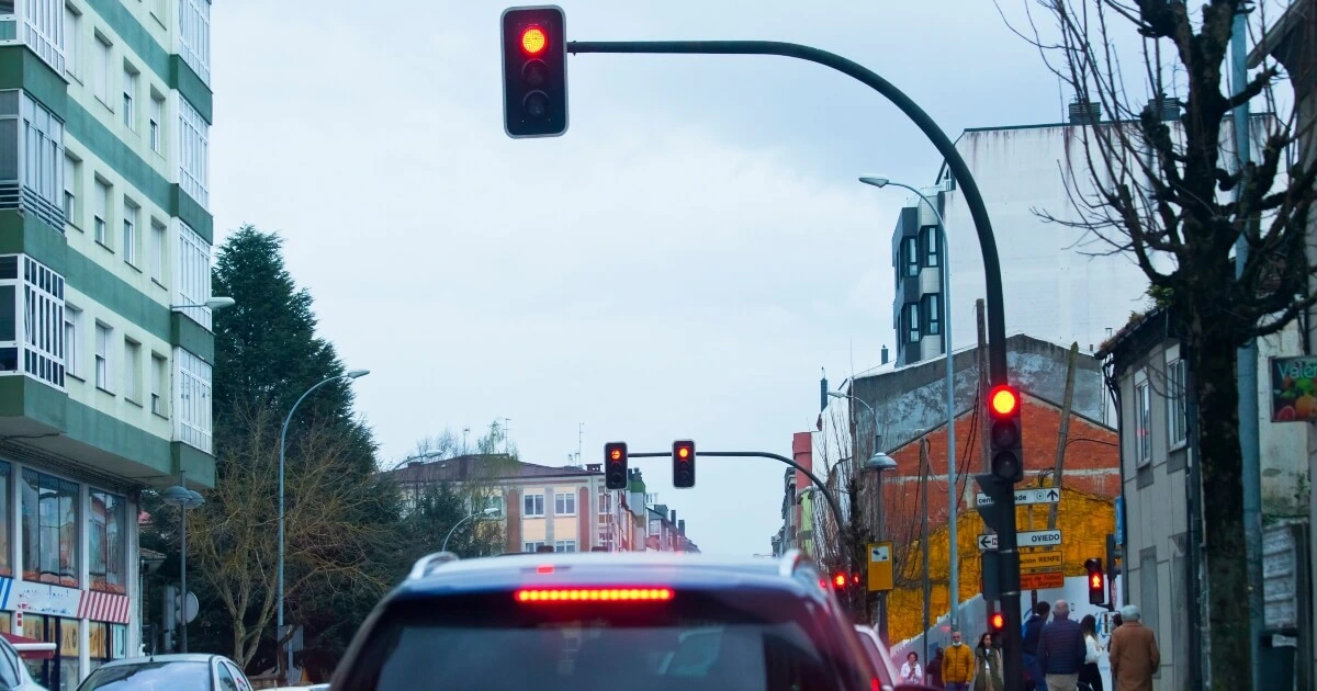 A city street scene viewed from behind a car at a red traffic light, with buildings, pedestrians, and cloudy sky in the background.