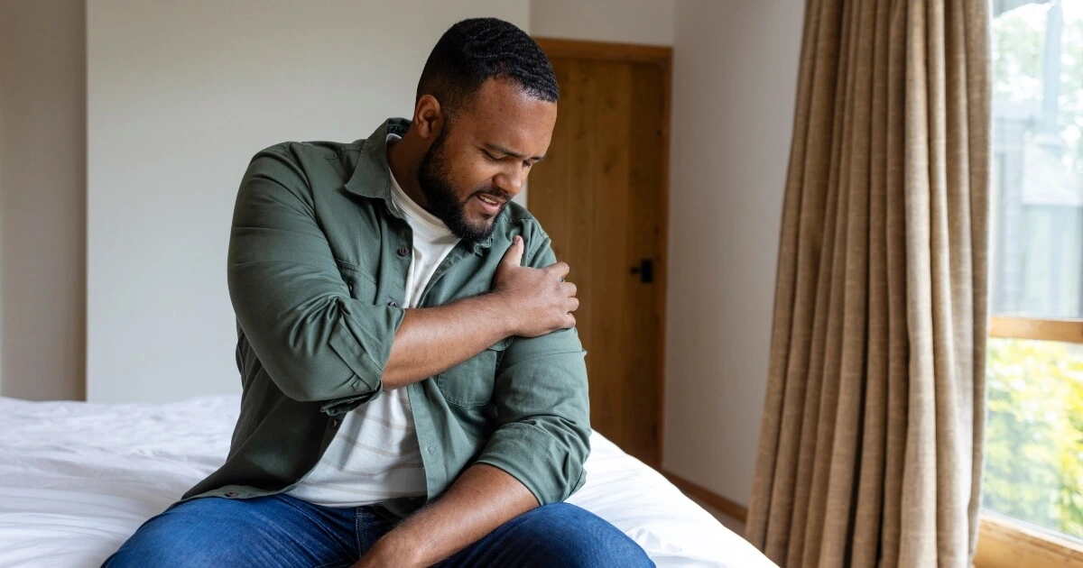 A man sits on a bed, holding his left shoulder and appearing to be in pain.