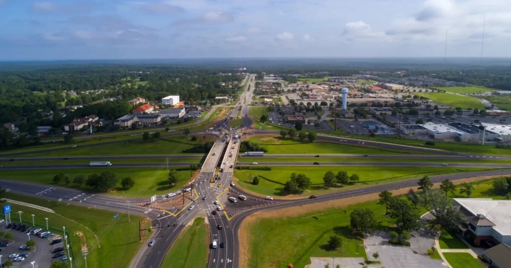 Aerial view of a diverging diamond interchange connecting two highways, surrounded by businesses, parking lots, and green areas under a partly cloudy sky.