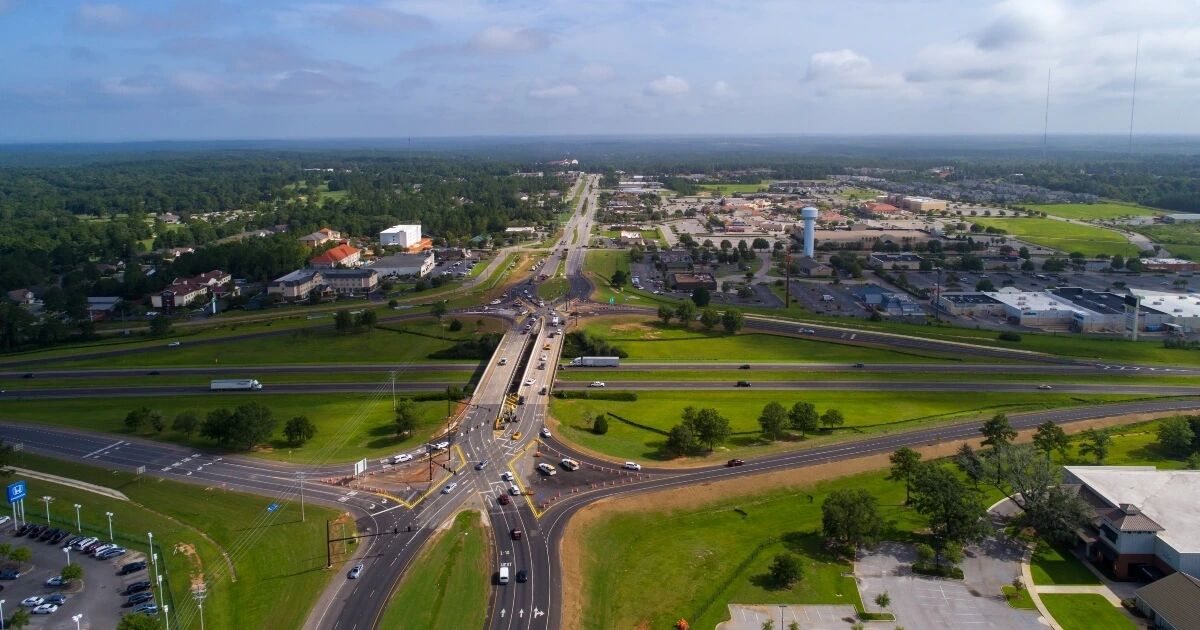Aerial view of a diverging diamond interchange connecting two highways, surrounded by businesses, parking lots, and green areas under a partly cloudy sky.