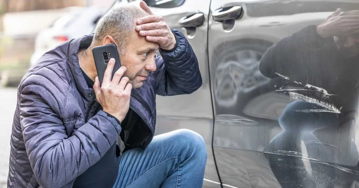 A man holding his head and talking on the phone examines scratches on the side of a gray car.