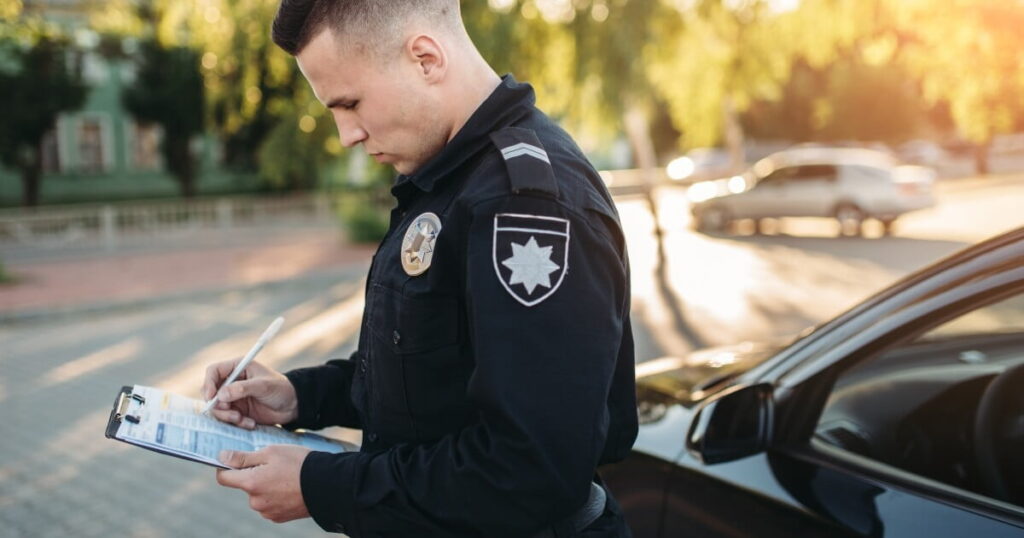 A police officer stands next to a car, writing on a clipboard in an outdoor urban setting during daylight.