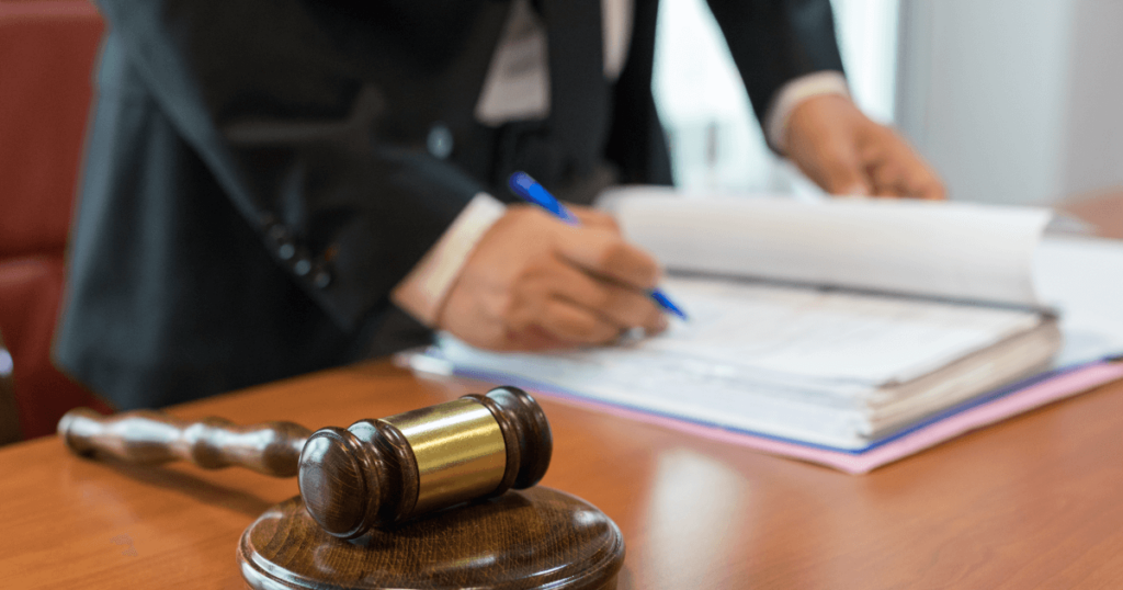 A person in a suit signs documents at a desk with a wooden gavel in the foreground.