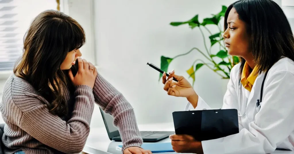 A doctor holding a pen and clipboard talks to a patient who appears concerned, sitting at a desk with a laptop.