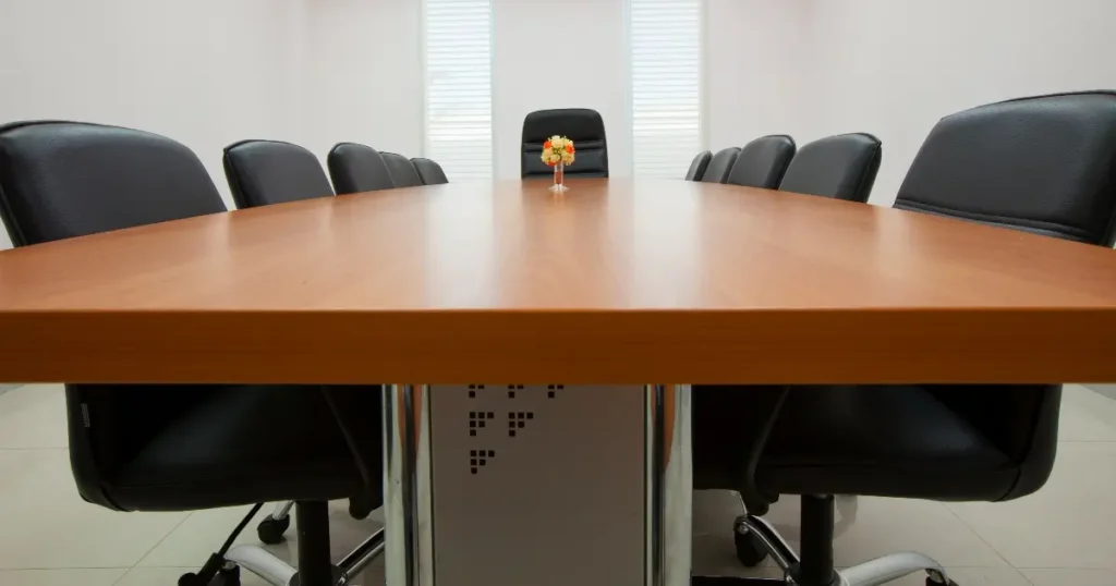 A conference room with a long wooden table, eight black office chairs, and a small flower arrangement in the center. Two windows with blinds are in the background.