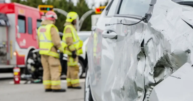 A close-up of a damaged silver car with two firefighters and a fire truck in the background at the scene of an accident.