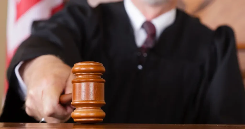 A judge in a black robe holds and strikes a wooden gavel on a desk, with a blurred flag visible in the background.