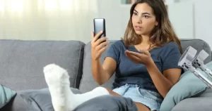 A woman with a leg cast sits on a couch, holding a smartphone and gesturing, with crutches beside her.