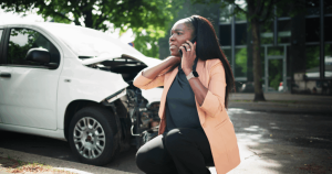 A woman kneels on the street while making a phone call, with a damaged white car and trees in the background.