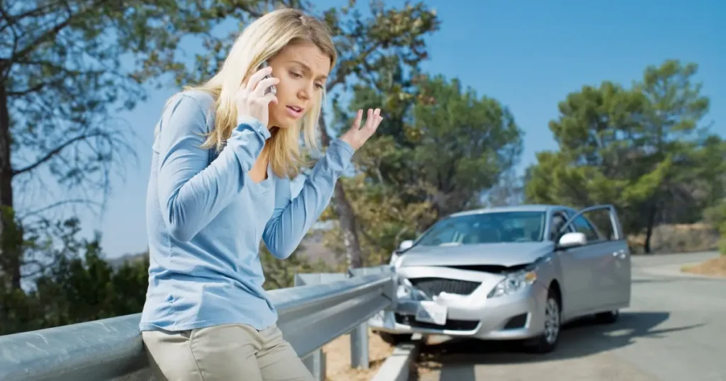 A woman stands by a guardrail on her phone, appearing distressed, with a damaged car in the background on a curved road.