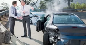 Two men stand on a road arguing next to two damaged cars after a collision, with smoke rising from one vehicle.
