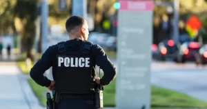 A police officer wearing a tactical vest labeled “POLICE” stands outdoors on a city street, facing away from the camera.