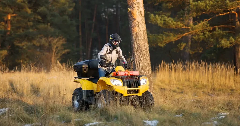 Person wearing a helmet and jacket rides a yellow ATV through a grassy field near trees on a sunny day.
