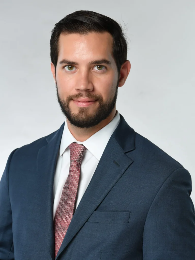 A man with dark hair and a beard wearing a blue suit, white shirt, and pink patterned tie poses against a plain light background.