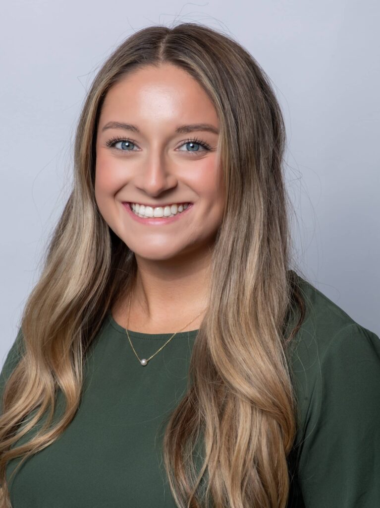 A woman with long, light brown hair and a green top smiles at the camera against a plain light gray background.