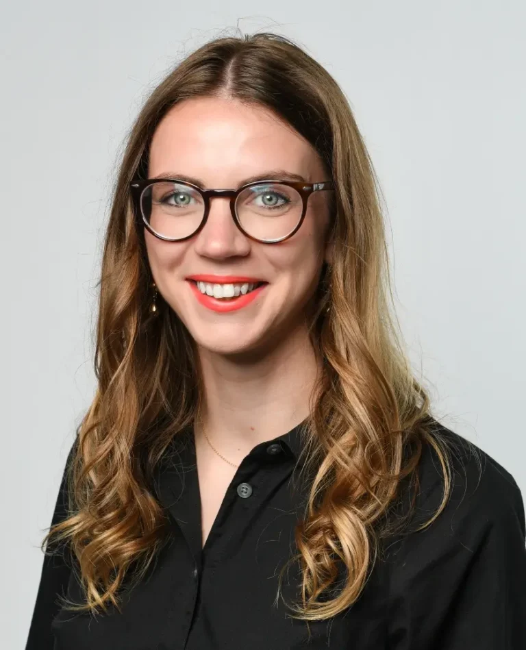 A woman with long, wavy, light brown hair and glasses is smiling at the camera. She is wearing a black shirt and has red lipstick. The background is plain light gray.