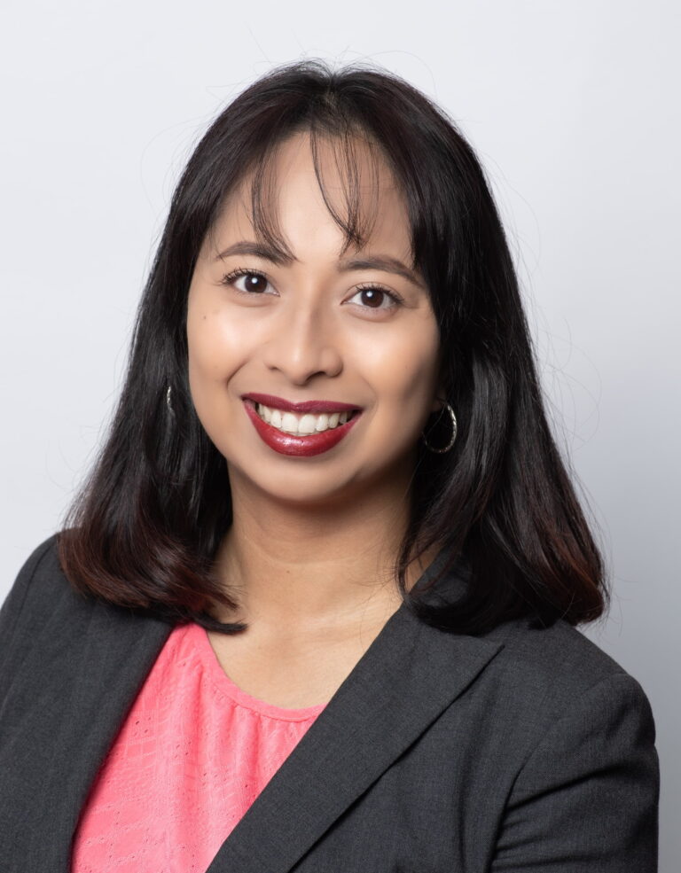 A woman with straight dark hair, wearing a dark blazer over a pink top, smiles at the camera against a plain light background.