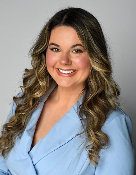 A woman with long wavy brown hair wearing a light blue blazer smiles at the camera against a plain gray background.