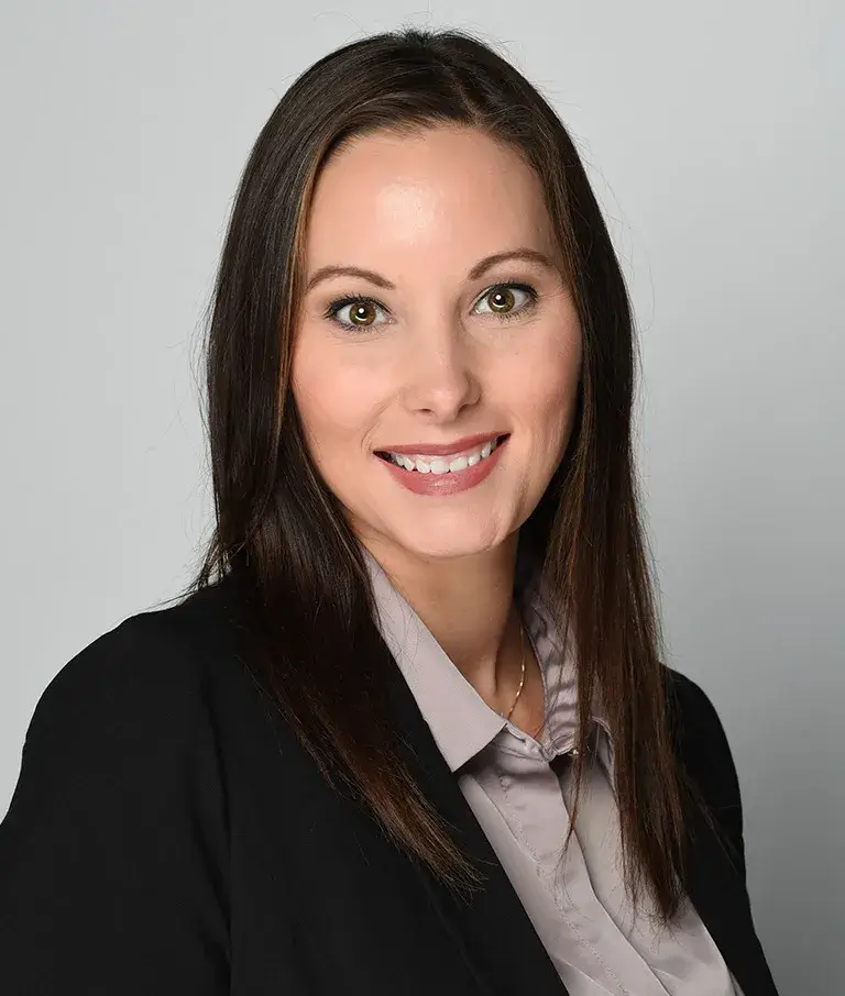 A woman with straight brown hair, wearing a black blazer and light-colored blouse, smiles at the camera against a plain light gray background.