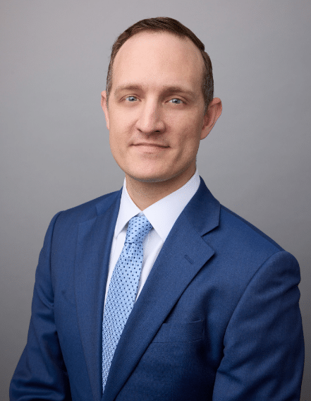 A man wearing a blue suit, white shirt, and light blue patterned tie poses for a professional portrait against a plain gray background.