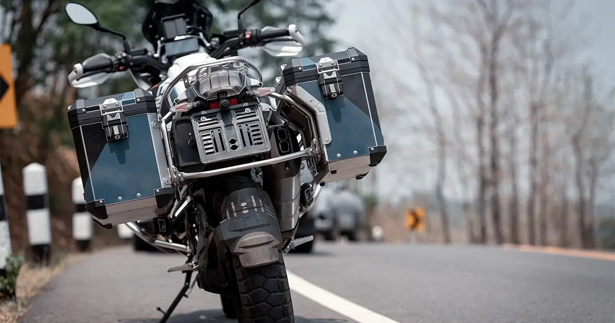 A touring motorcycle with saddlebags is parked on the side of a paved road with trees and road signs visible in the background.
