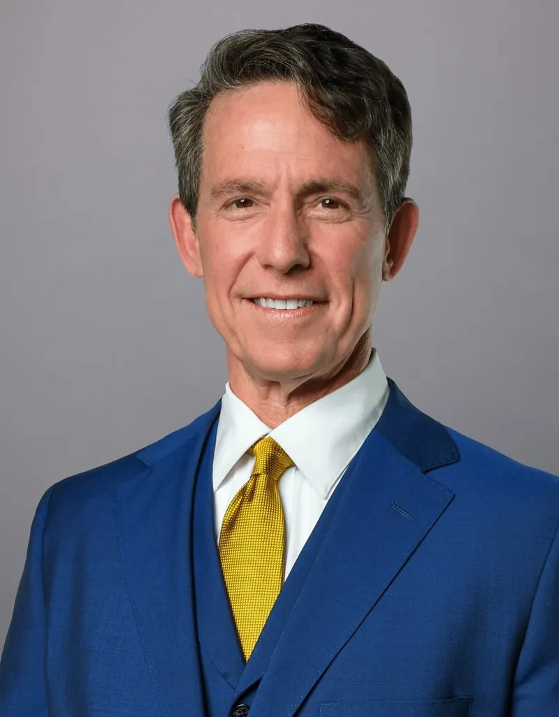 A man wearing a blue suit, white dress shirt, and yellow tie poses in front of a plain gray background, smiling at the camera.