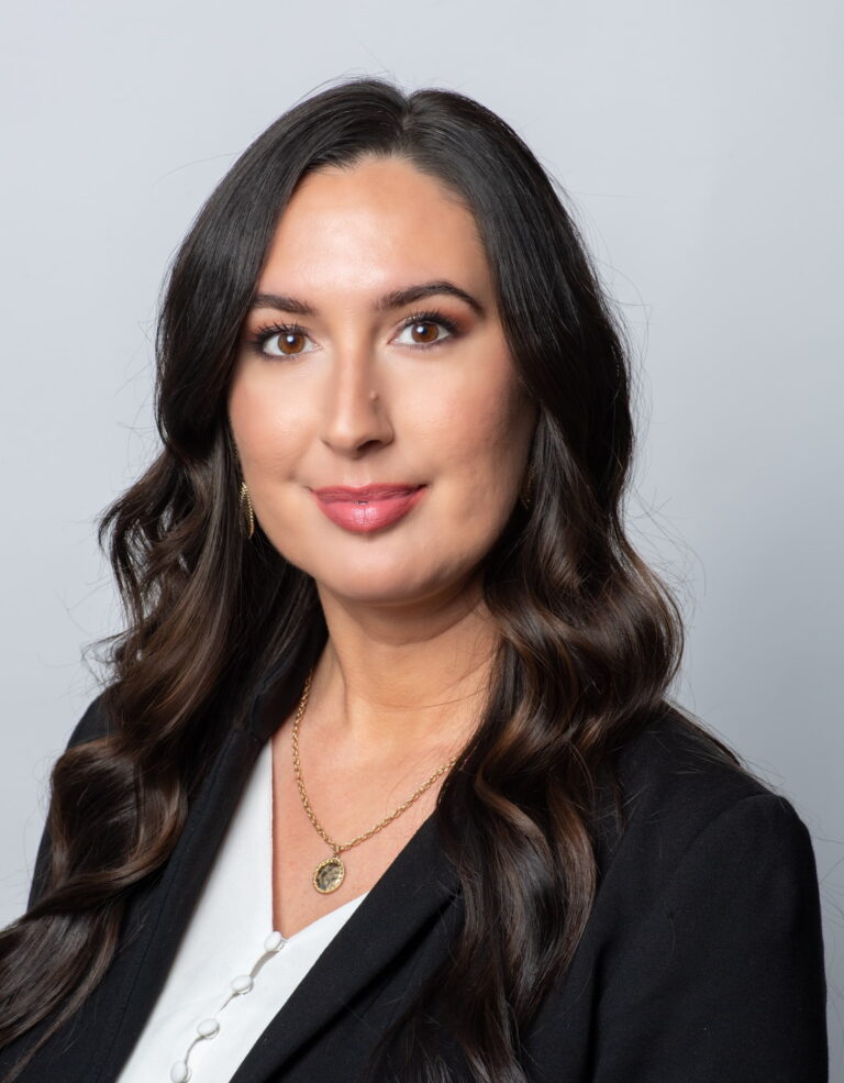 Woman with long dark hair wearing a black blazer and white top, facing the camera with a neutral expression against a plain light background.