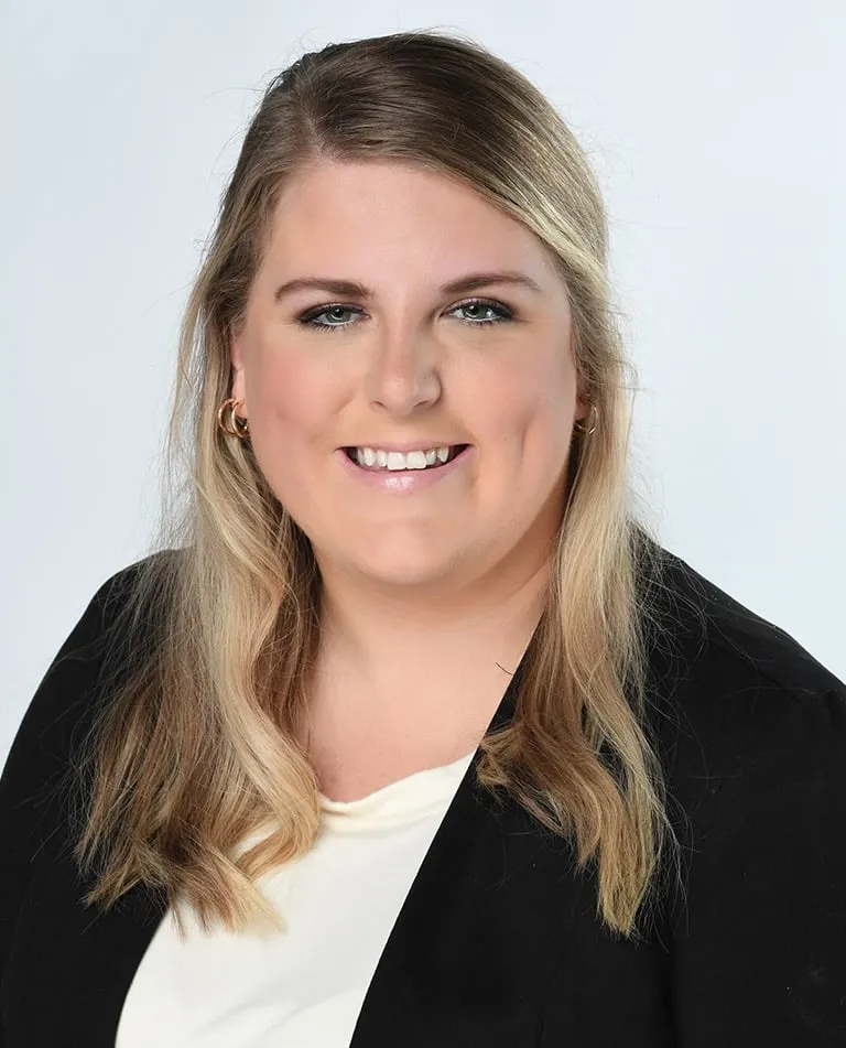 A woman with long blonde hair wearing a black blazer and white top smiles at the camera against a plain light background.