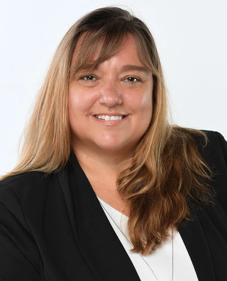 A woman with long, light brown hair wearing a black blazer and white top, smiling at the camera against a plain white background.