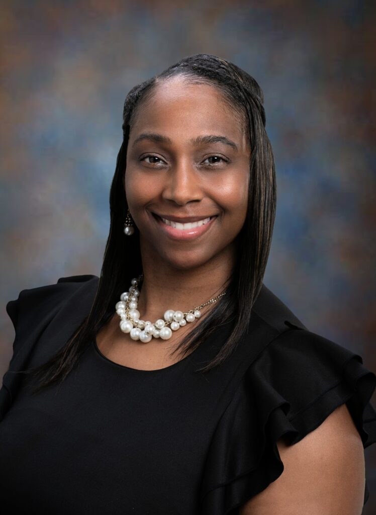 Woman with straight dark hair, wearing a black dress, pearl necklace, and earrings, smiling in front of a neutral, softly blurred background.